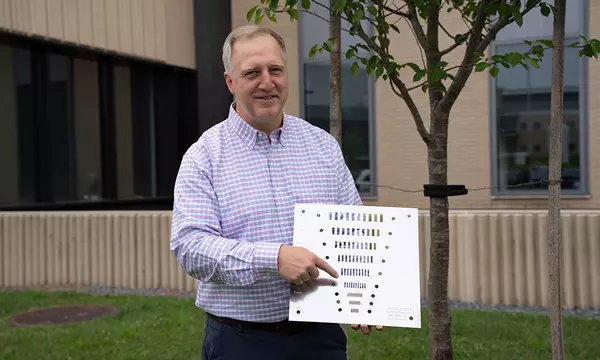Jack Glover poses outside on the NIST campus, holding a flat square object with holes of varying sizes. 