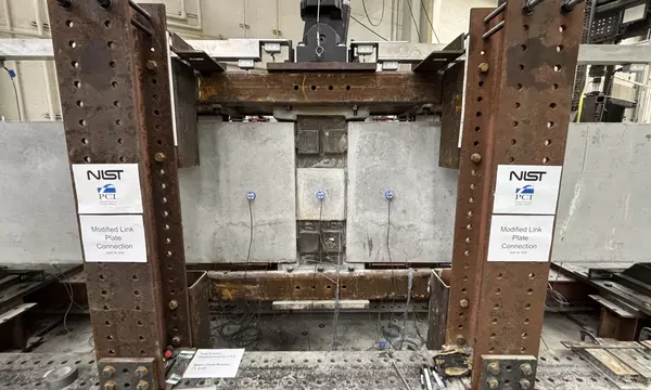 A wide view of a concrete beam assembly inside a large metal frame. A hydraulic arm is part of the metal frame, and it’s posed to press down at a section in the middle where the concrete pieces connect.
