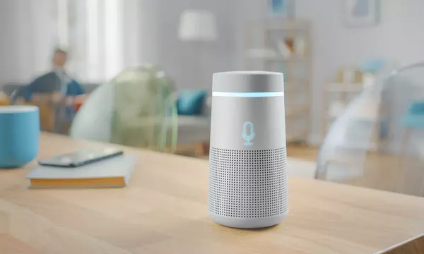 A cylindrical smart speaker sits on a wooden table in a living room. 