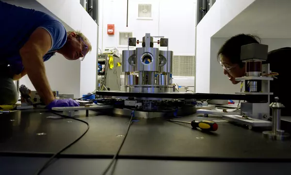 Wide shot shows two researchers on either side of a scientific device made of metal cylinders, peering closely at it. 