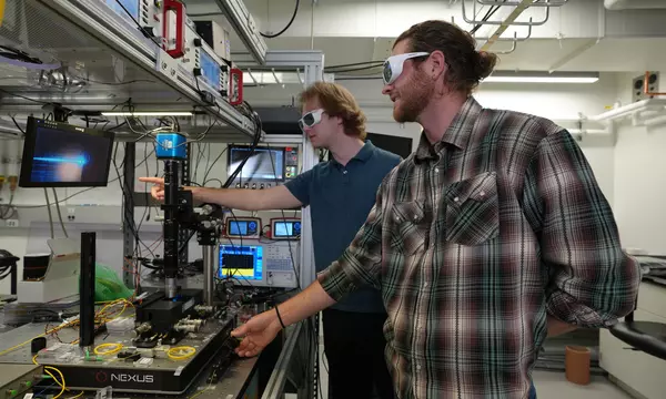 Two male researchers in casual clothes and protective eyewear stand in the lab, pointing at a screen hooked up to a laser science instrument. 