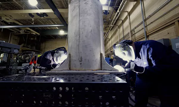 Two people wearing welding masks and other safety equipment bend over a table to work on a large metal cylinder.