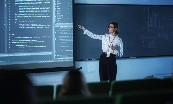 A female instructor wearing glasses presents code on a large screen to a classroom.