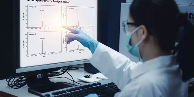 A researcher wearing a mask and gloves points at a computer screen showing graphs labeled "Mass Spectrometry Analysis Report."