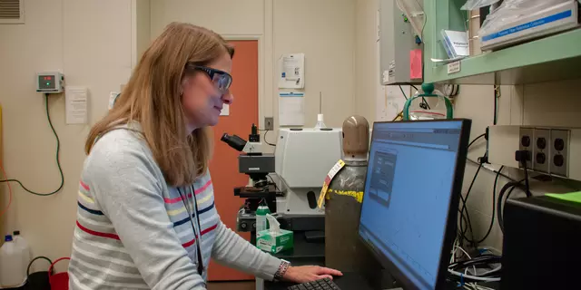 A scientist wearing safety glasses stands in front of a monitor in her lab.