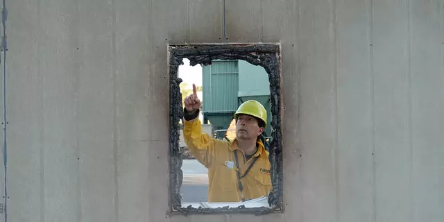 A man wearing a yellow hardhat and fire-resistant jacket points to burnt frame of a window.