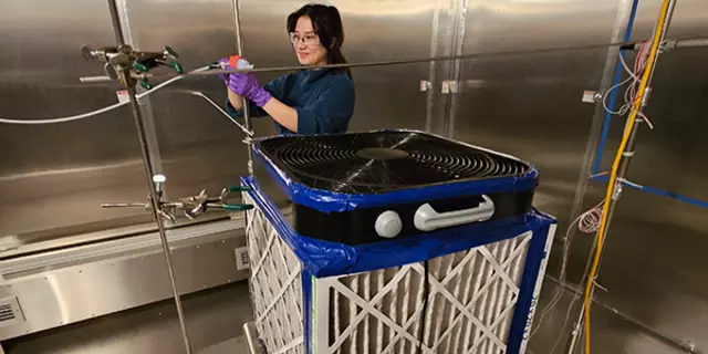 A large cube made from a box fan, pleated air filters, and duct tape sits on a table in the middle of a large air tight steel room. In the background, a scientist clamps a narrow, flexible hose in place.