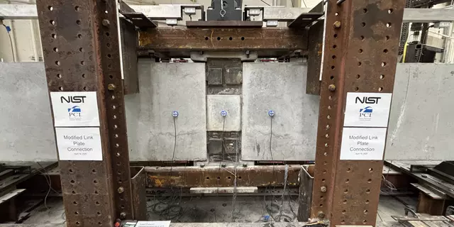 A wide view of a concrete beam assembly inside a large metal frame. A hydraulic arm is part of the metal frame, and it’s posed to press down at a section in the middle where the concrete pieces connect.