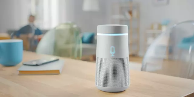 A cylindrical smart speaker sits on a wooden table in a living room. 
