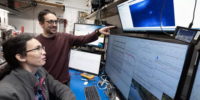 A woman with glasses and a long ponytail is seated at a computer with four display screens. A man with glasses stands beside her and points at the uppermost display screen. In the middle of that screen is a small blue glowing dot. The lower screens display data from the experiment.