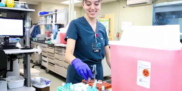 A medical worker in scrubs places a vial into a rack in a medical lab. 