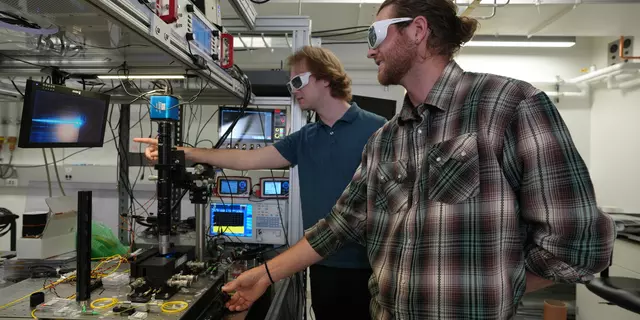 Two male researchers in casual clothes and protective eyewear stand in the lab, pointing at a screen hooked up to a laser science instrument. 