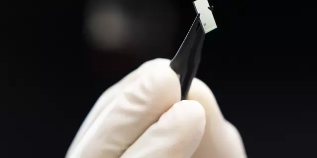 Close-up of a gloved hand holding tweezers that are grasping a tiny rectangular chip. 
