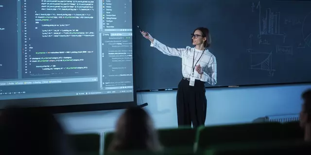 A female instructor wearing glasses presents code on a large screen to a classroom.