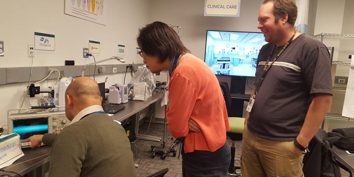 One person sits in front of a wireless infusion pump. Two people stand and look over his shoulder. Other pumps are in the background.