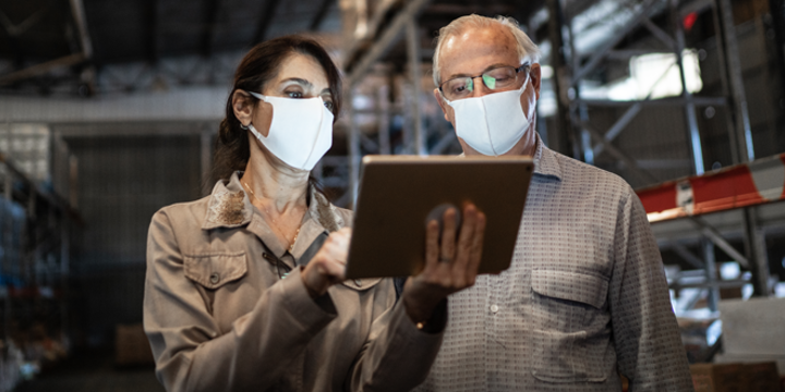 Senior partners walking and using digital tablet at warehouse with face masks