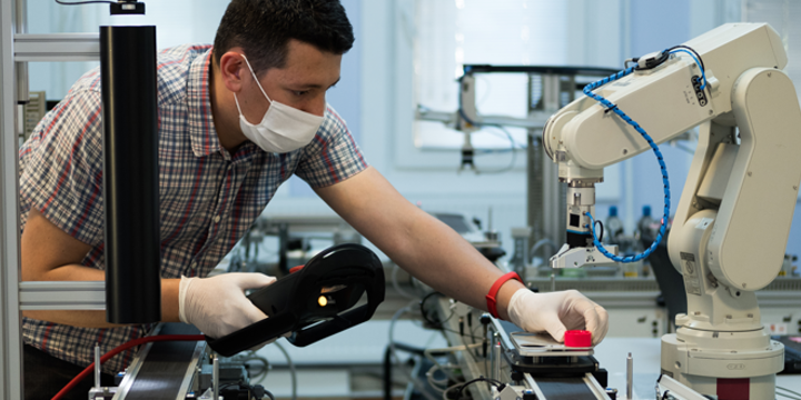 Man with surgical mask and gloves against COVID-19 ( Coronavirus ) programming robot arm with teach pendant.