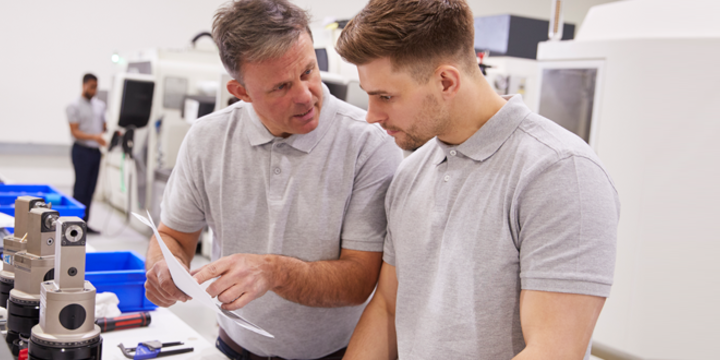 Engineer And Apprentice Discussing Job Sheet In Factory 