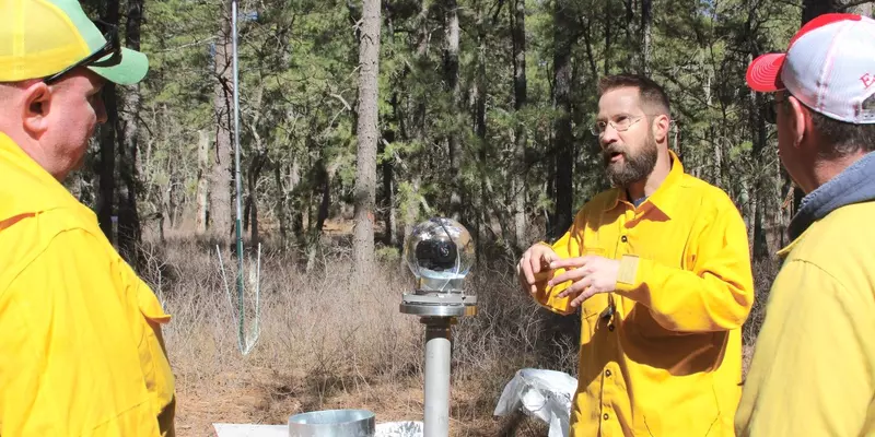 NIST engineer in yellow jacket stands to right of BOB glass globe. To his right and left are two men.