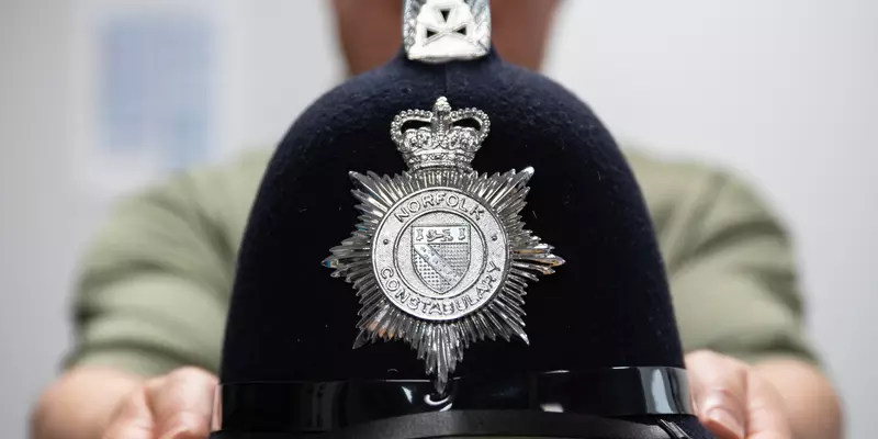 A blurry figure in the background holds out a black police helmet with a silver seal that reads "Norfolk Constabulary."