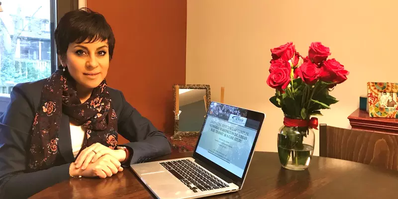 Yalda Saadat poses sitting at a table at home, next to an open laptop and a vase of roses. 
