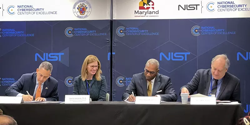 Four officials sit at a table signing documents in front of banners for the NIST National Cybersecurity Center of Excellence.