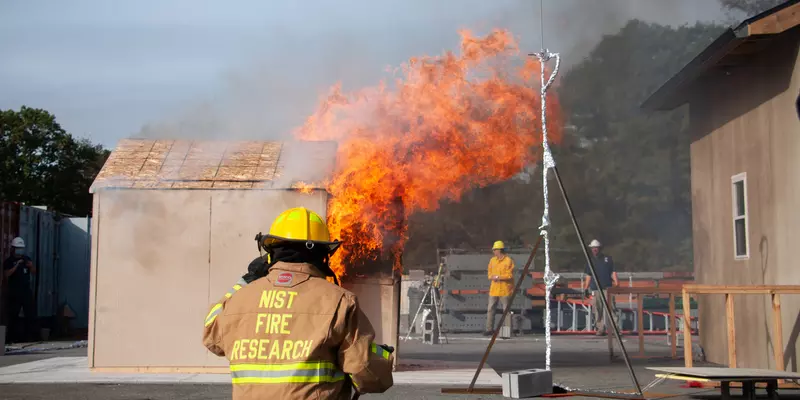 Photo shows a NIST firefighter on standby watching a shed burn next to a target structure representing a wall of a residence during an experiment.