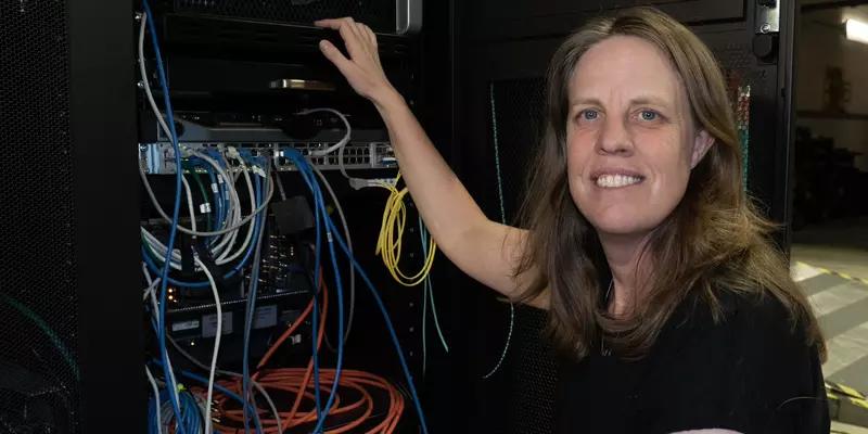 Jeanne Quimby crouches next to a rack of computer equipment with many different colored cord connections. 