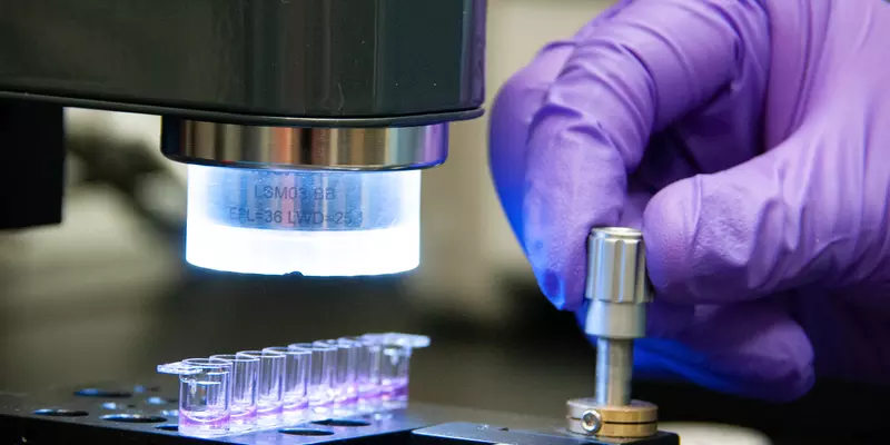 A hand in a purple glove turns a metal dial on a plate holding tiny vials of purple liquid. 