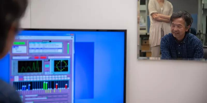 A mirror reflects Yoshi Ohno, seated, and Jane Li, standing, as they view a computer screen showing lighting data. 