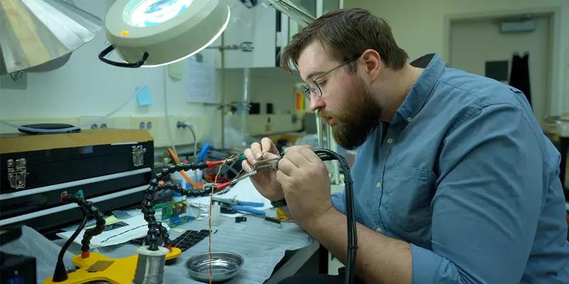Peter Trask sits at a lab table working on wiring for a scientific device. 