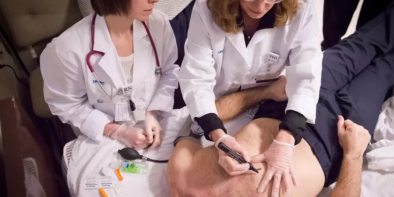 Two nurses sit next to a bed where a shirtless man lies on his back. One nurse uses a marker to make an X on the man's chest. 