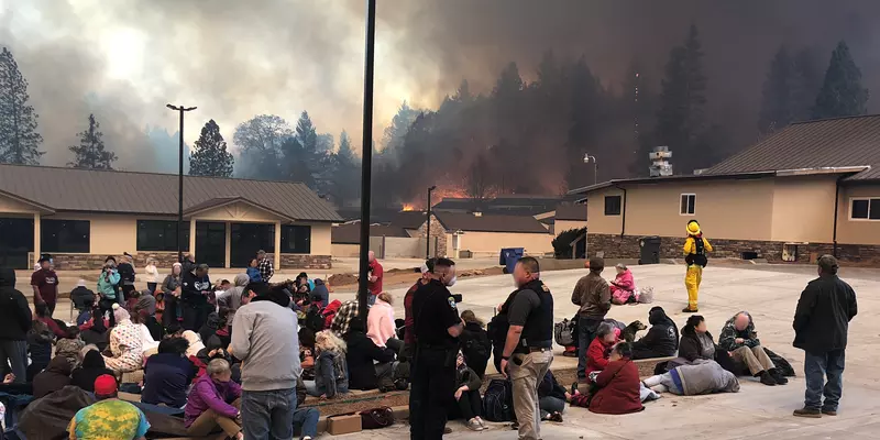 Groups of people sit or lie in a parking lot surrounded by low buildings as fires burn in the forest beyond.  