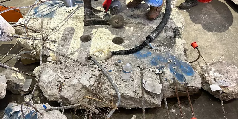 A slab of concrete with ragged edges sits on a warehouse floor as researchers use equipment to extract core samples. 