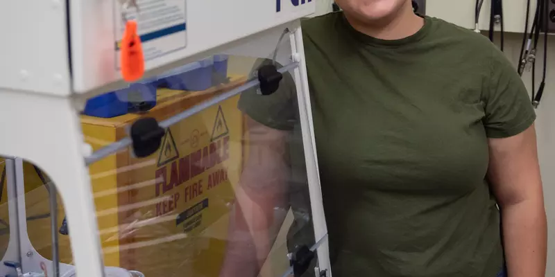 Woman in safety glasses stands next to an enclosed space with scientific equipment inside.