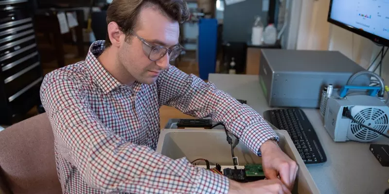 A man wearing safety glasses and sitting at a computer workstation reaches into a box with wiring and circuitry.