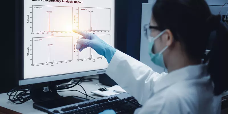 A researcher wearing a mask and gloves points at a computer screen showing graphs labeled "Mass Spectrometry Analysis Report."