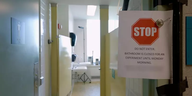 A paper sign is clipped in front of a open door to a public restroom. The sign reads, “Stop. Do Not Enter. Bathroom is closed for an experiment until Monday Morning.”
