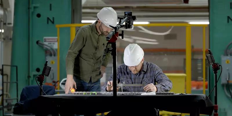 Two researchers in hard hats lean over a rebar sample on a table in a large workspace. 