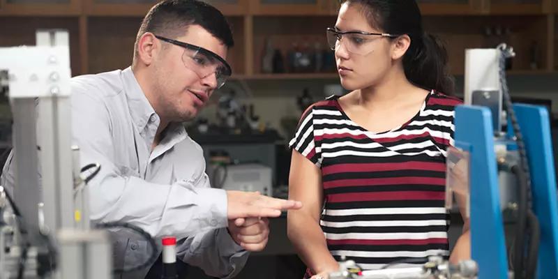 A man wearing safety glasses gestures as he explains something to a woman watching attentively, in a lab. 