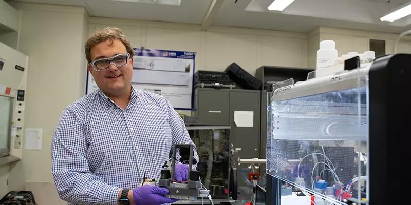 Peter Beaucage wears safety glasses as he stands in the lab, holding a piece of equipment on display. 