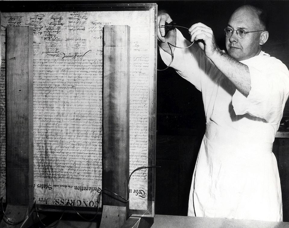 A NIST scientist stands before the Declaration of Independence, which is upright in glass, and works on the encasement