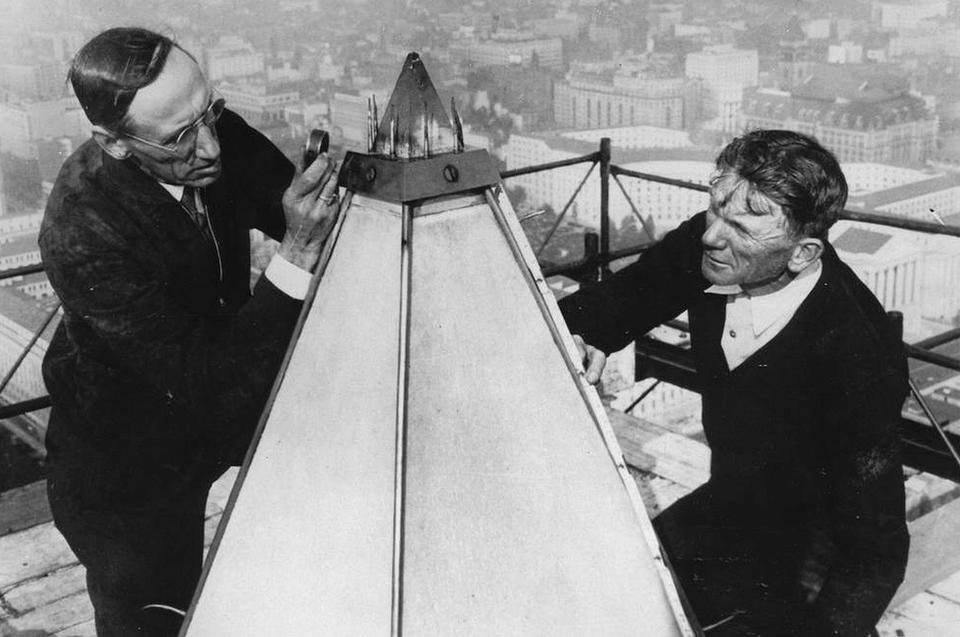 Two men sit on scaffolding and examine the tip of the Washington Monument.
