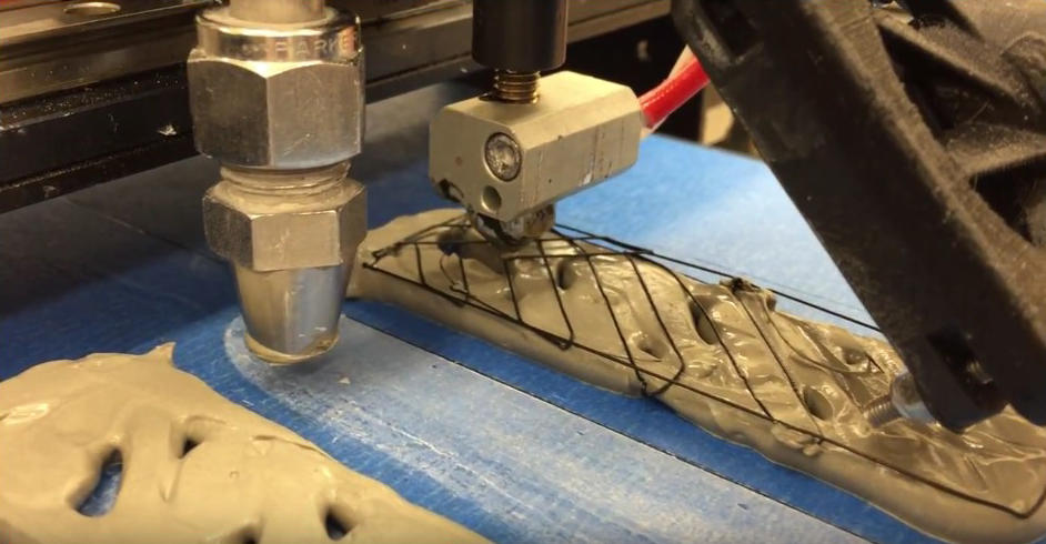 rectangle of tan-colored concrete on a platform. A 3D printer is laying down a layer of carbon fiber filament on top of the concrete. 