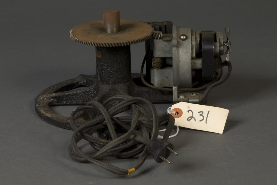 geared mechanism with an electric plug. Attached to the plug's wire is a tag that reads "231." The device is geared to another smaller gear on a device with what looks like a flywheel coming from the front of it. 