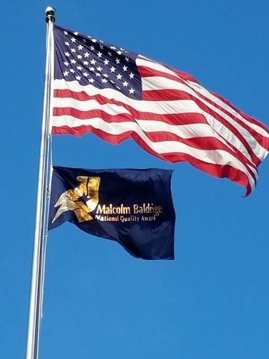 Baldrige program flag and American flag waving on steel pole against blue sky