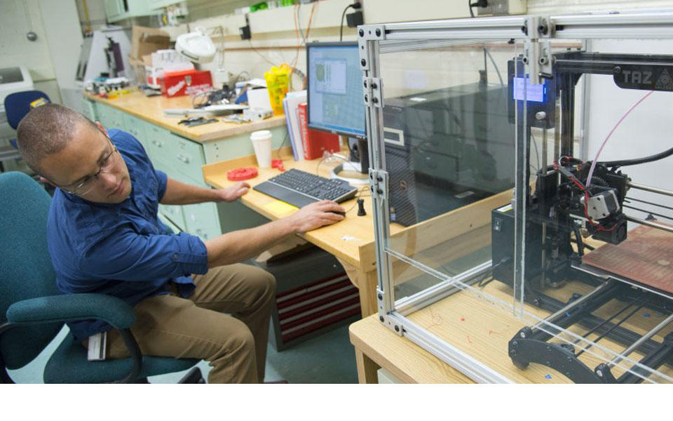 Man in blue shirt working at computer looking at device in lab