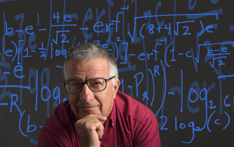 Former NIST researcher Tassos Nakassis sitting in front of a black background. The background is covered in blue equations.