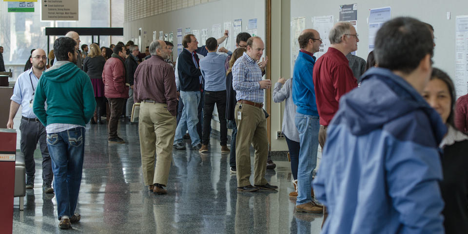 Scientific posters line a hallway. NIST postdocs and researchers stand in front of the posters discussing them.