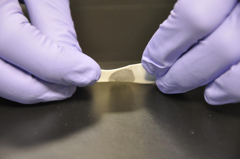 A close up of two purple gloved hands removing a piece of tape from a shiny surface. A fingerprint can be seen on the tape.